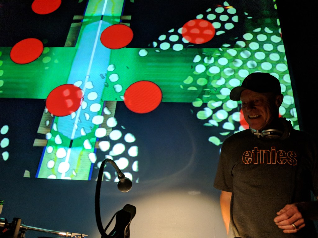 Rich Thair from Red Snapper stands in front of record decks with a projection screen behind him filled with texture, and red dots.