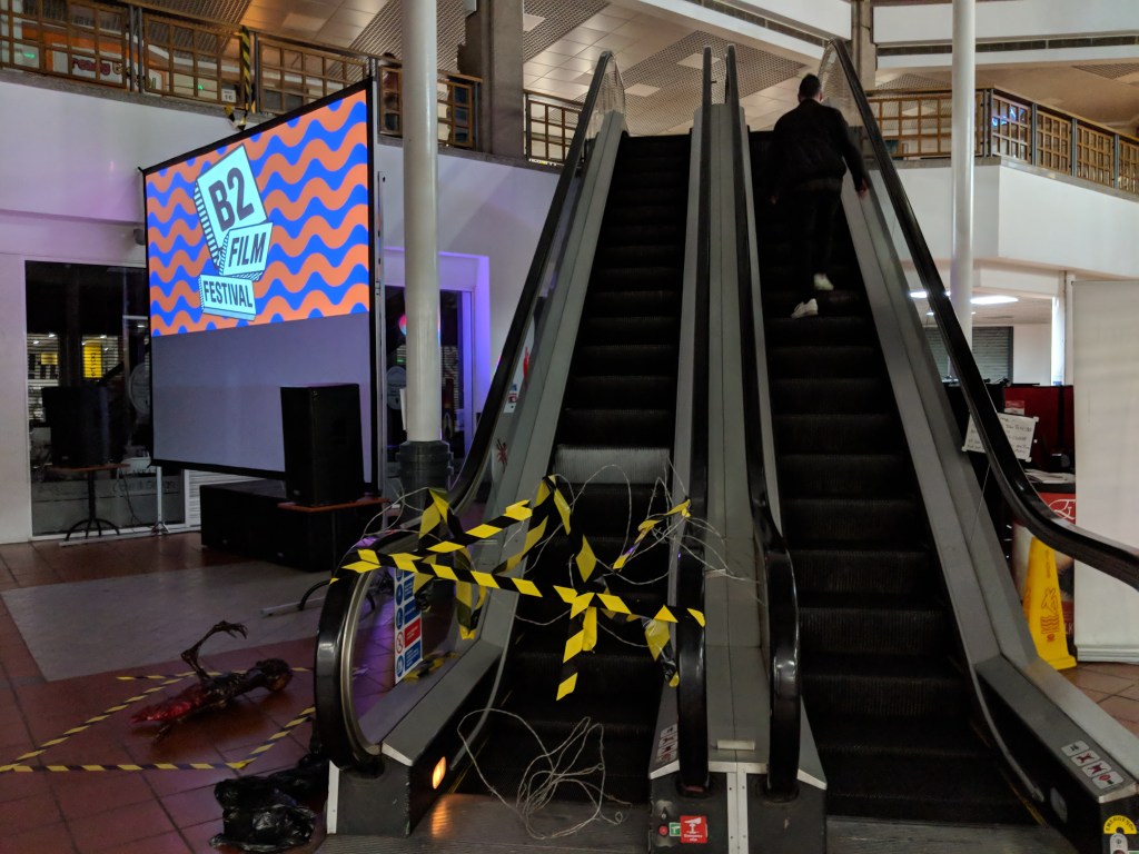 Someone is walking up a double escalator. The bottom of one of the escalators has hazard tape and barbed wire across it. There's a burnt corpse on the floor surrounded by hazard tape. A large projection screen has the title 'B2 Film Festival' projected on to it.