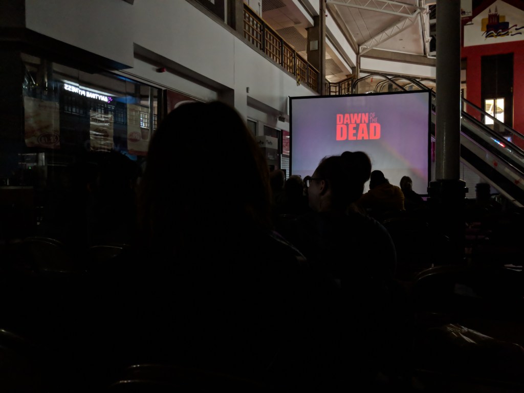 A crowd sit in a dark shopping mall staring at a large screen which has the title ' Dawn of the Dead' projected on to it